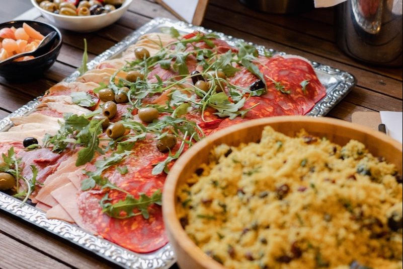 A platter of charcuterie and salad displayed on a wooden table at The Square Club, a premium party venue in Bristol. The platter includes thinly sliced meats like salami and prosciutto, garnished with arugula and green olives. Next to it is a large bowl of couscous mixed with raisins and herbs. This sophisticated and delicious spread highlights the catering quality for events and celebrations held at this renowned party venue in Bristol.