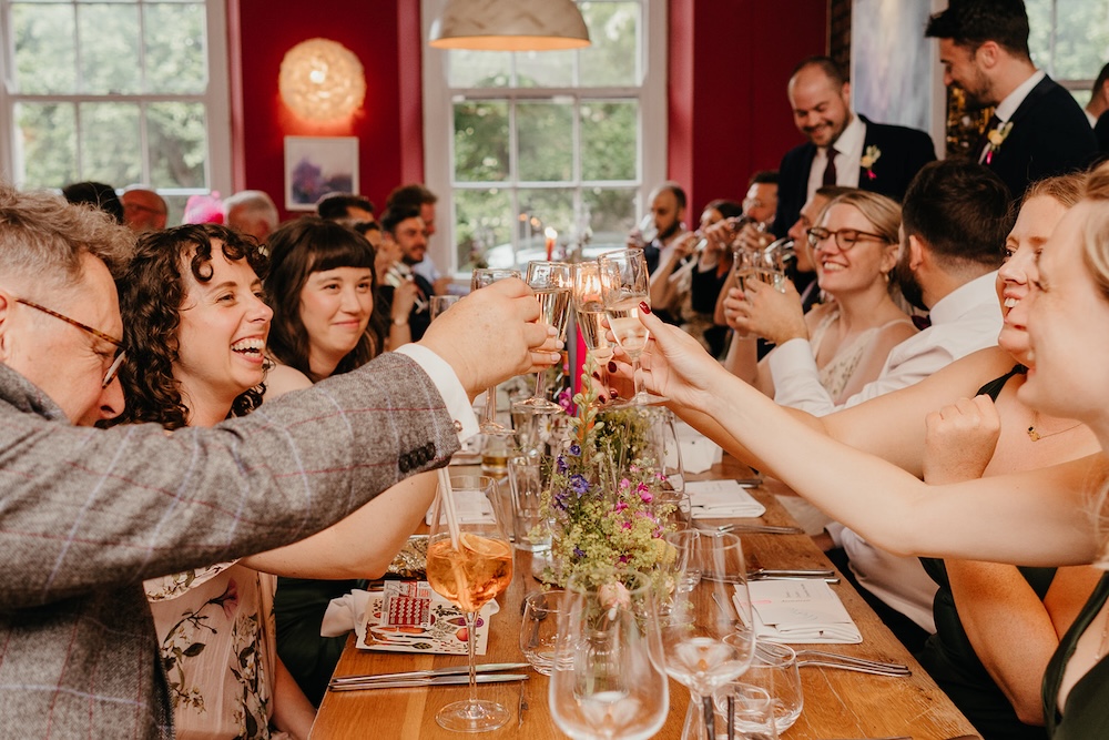 A large group of guests raising glasses in a celebratory toast at a wedding reception held at The Square Club, a premier party venue in Bristol. The long dining table is decorated with floral arrangements and elegant glassware. Guests are smiling and laughing, capturing the joyful atmosphere of the event. This image showcases the venue’s ability to host large-scale celebrations, from weddings to corporate events, with a lively and festive vibe.