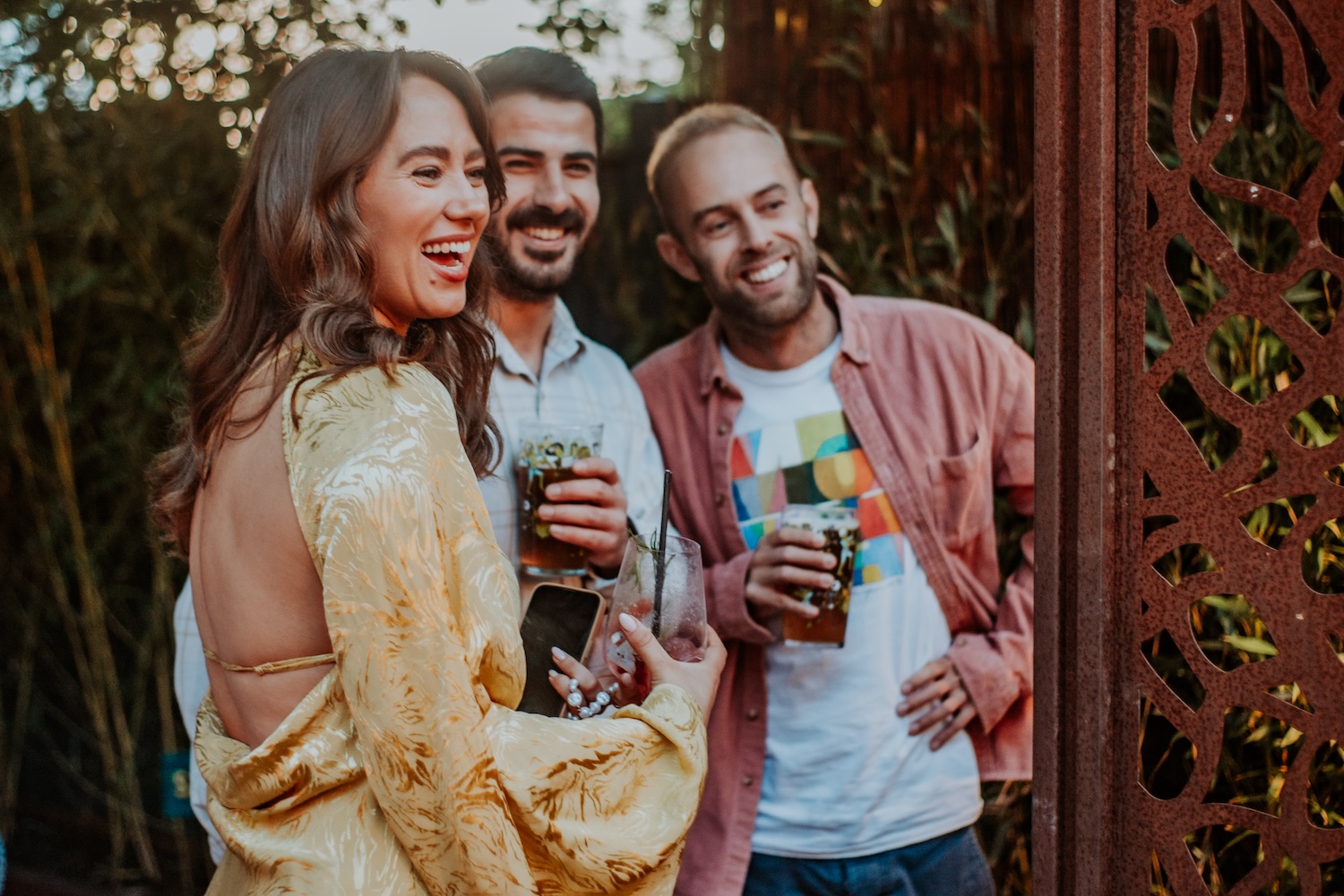 Three friends enjoying drinks and laughter at an outdoor party venue in Bristol, hosted at The Square Club. A woman in a shiny gold dress is turned toward the camera, while two men stand behind her, holding glasses filled with beer or cocktails. The scene captures a moment of joy, with the greenery and intricate wood paneling in the background adding to the venue’s stylish ambiance, making The Square Club a perfect location for private parties in Bristol.