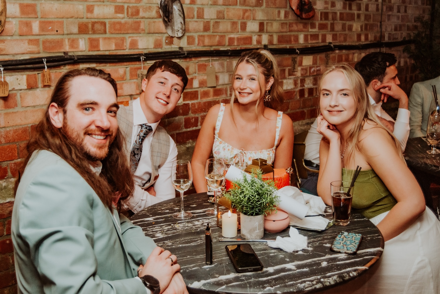 A group of four friends sitting at a round table inside The Square Club, a notable party venue in Bristol. The group, consisting of two men and two women, are dressed stylishly, enjoying their drinks and conversation. The setting is intimate, with a brick wall in the background and warm, ambient lighting, making this a perfect snapshot of a relaxed, social event at one of the best private party venues in Bristol.