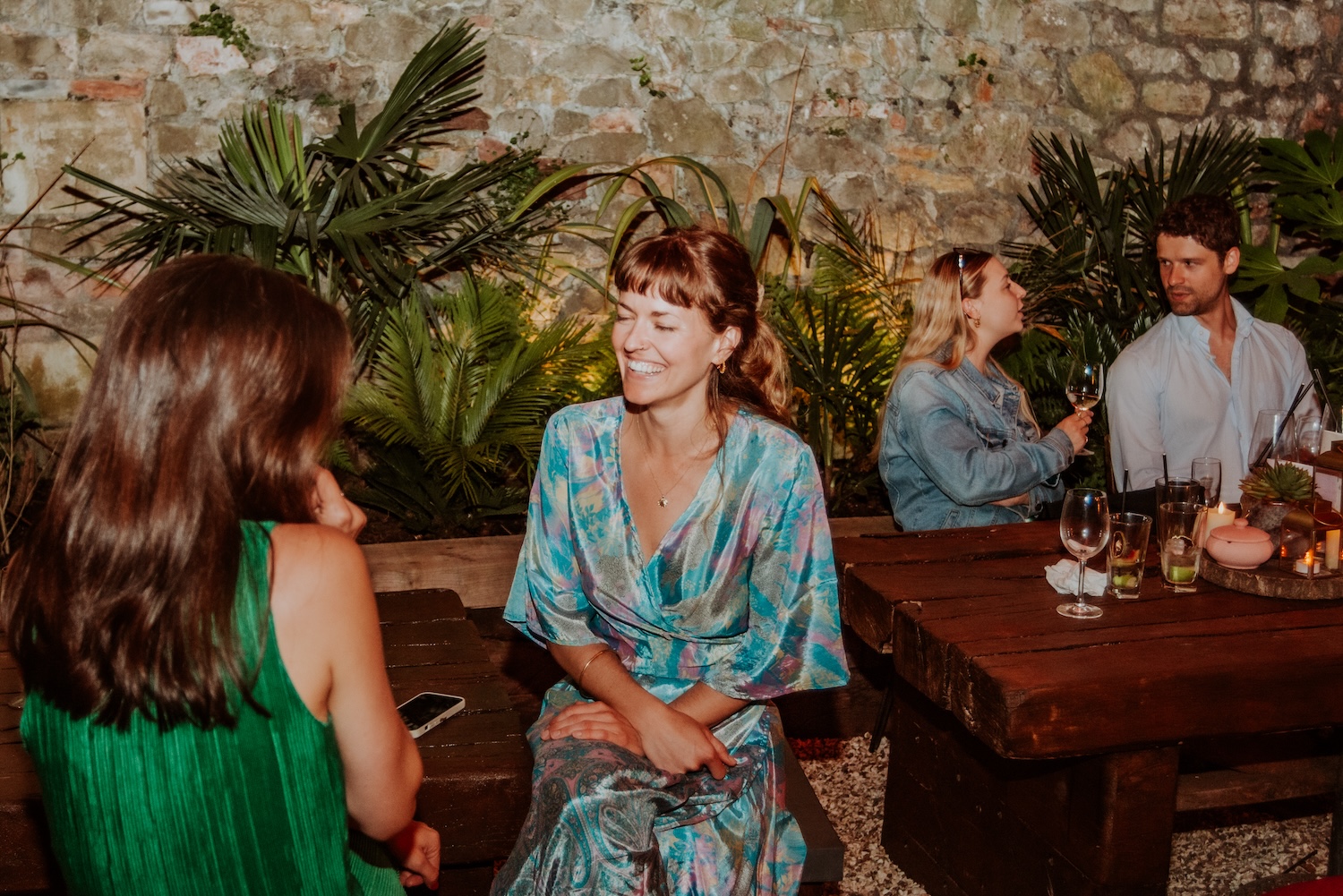 Two women engaged in conversation at an outdoor party venue in Bristol, The Square Club. One woman, in a green pleated dress, faces away from the camera, while the other, seated at a wooden bench in a colorful wrap dress, smiles with her eyes closed. The lush greenery surrounding them and the rustic stone wall create a serene atmosphere, showcasing The Square Club's versatility for both lively and intimate gatherings in Bristol.