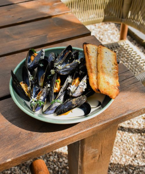 A close-up of a dish featuring steamed mussels served in a light creamy broth, garnished with finely chopped herbs and edible flowers. The black mussel shells contrast with the bright orange flesh inside, creating a vibrant and appetizing presentation. Toasted bread is served on the side for dipping into the sauce. This seafood dish, likely from a restaurant in Clifton, captures the essence of fine dining with fresh ingredients. The setting suggests it could be a menu item from The Square Club, a restaurant in Bristol known for its refined cuisine and stylish ambiance.