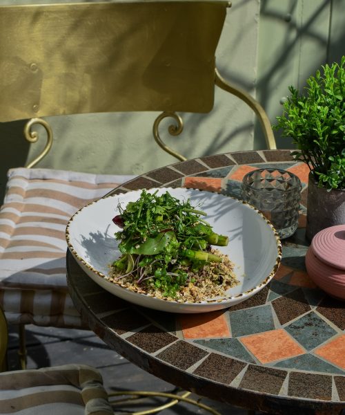 An outdoor table at a restaurant in Clifton features two elegant dishes: a plate of fish and chips with a side of mushy peas and a light, vibrant salad with fresh greens and quinoa. The table is positioned in a serene garden setting, suggesting a quiet and inviting atmosphere at The Square Club, a stylish restaurant in Bristol. The dappled sunlight enhances the natural feel of this dining experience, making it a perfect representation of a relaxing meal in one of the city’s hidden culinary gems.