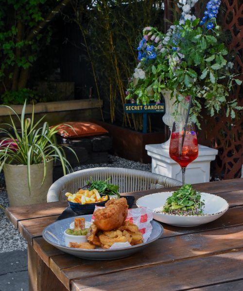 A rustic outdoor dining setup at a restaurant in Clifton shows a wooden table adorned with two plates of food, one featuring a classic fish and chips dish, and the other a fresh salad. The setting, shaded by greenery and complemented by a glass of red aperitif, suggests an idyllic, tranquil atmosphere perfect for leisurely dining. A sign reading "Secret Garden" is visible in the background, hinting at a hidden dining gem within The Square Club, a popular restaurant in Bristol known for its cozy garden space and artisanal dishes.