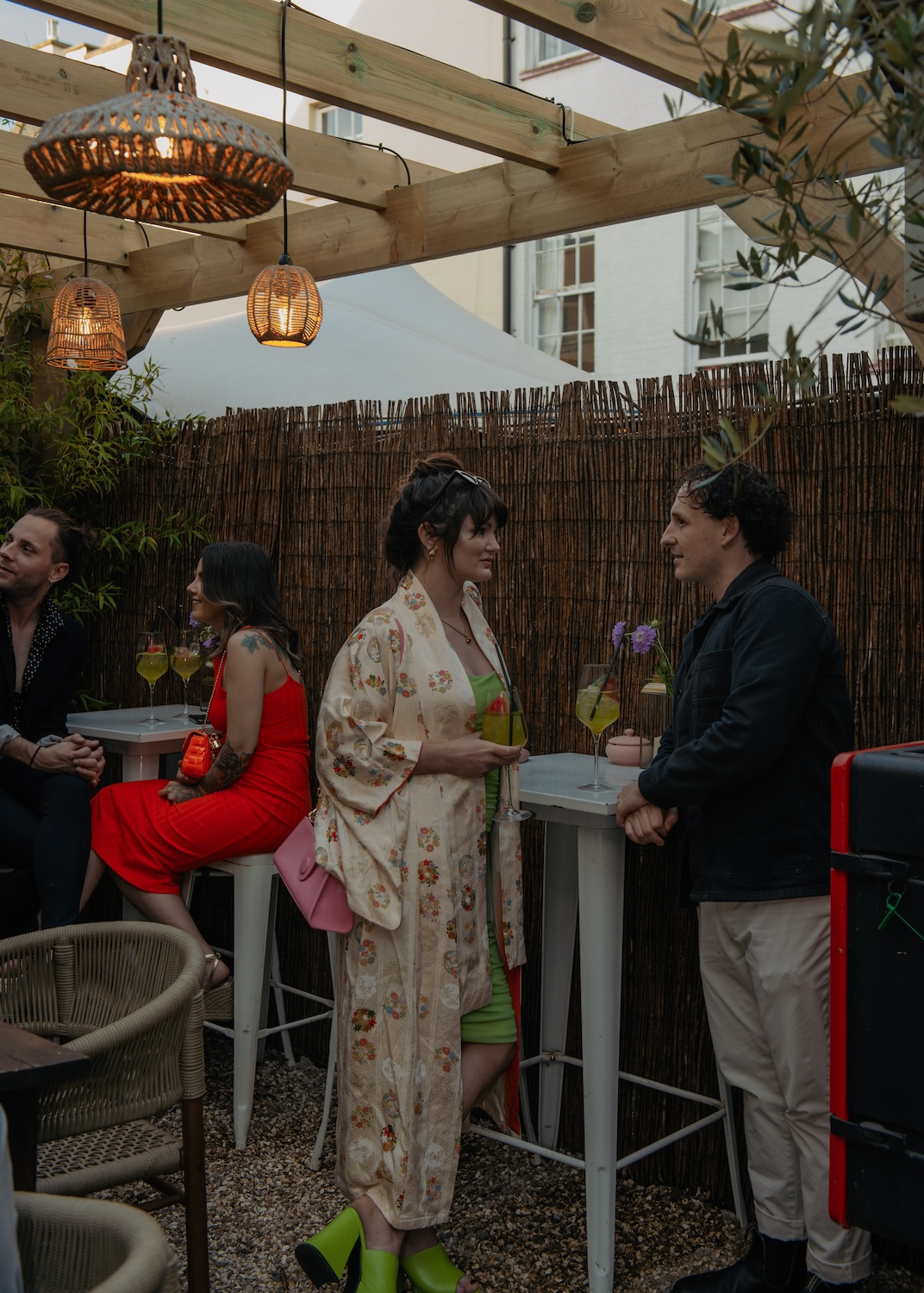 A group of people, including a woman in a floral kimono and bright green heels, are chatting at a rooftop party venue in Bristol, known as The Square Club. They are standing around high-top tables, sipping colorful drinks under the warm glow of wicker light fixtures. The venue's cozy, outdoor setting and stylish décor make it a go-to location for parties and casual gatherings in the heart of Bristol.