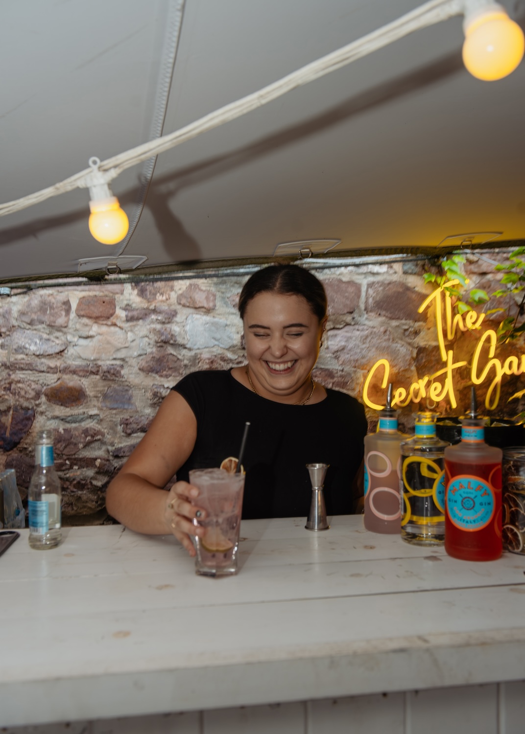 A bartender at The Square Club, a popular party venue in Bristol, smiles warmly as she prepares a cocktail. Behind her, the neon sign reads "The Secret Garden," adding a touch of elegance to the stone wall backdrop. The bar is decorated with premium gin bottles, and overhead lights create a cozy ambiance. This image highlights the intimate and stylish bar service provided for private events and celebrations at The Square Club, one of the best venues for hosting social gatherings in Bristol.