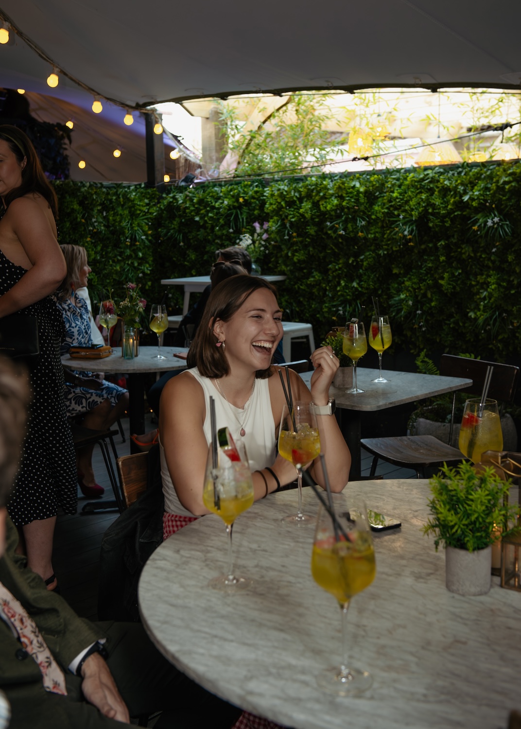 A woman sitting at an outdoor table at The Square Club, a prime party venue in Bristol, enjoys her drink while laughing with friends. The table is filled with cocktails garnished with fresh fruit, and the lush greenery surrounding the area creates a tranquil atmosphere. Twinkling string lights hang overhead, enhancing the venue's charm, which makes it ideal for any private party or social gathering in Bristol.