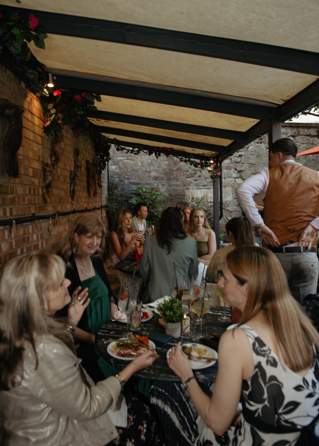 A lively gathering of people sitting under a canopy at The Square Club, one of Bristol’s best party venues. The group is seated at long tables, enjoying their food and drinks in a partially covered outdoor space. The brick and stone walls, along with the floral decorations, add to the intimate and welcoming vibe. The relaxed atmosphere makes this venue a top choice for private parties and dining events in Bristol.