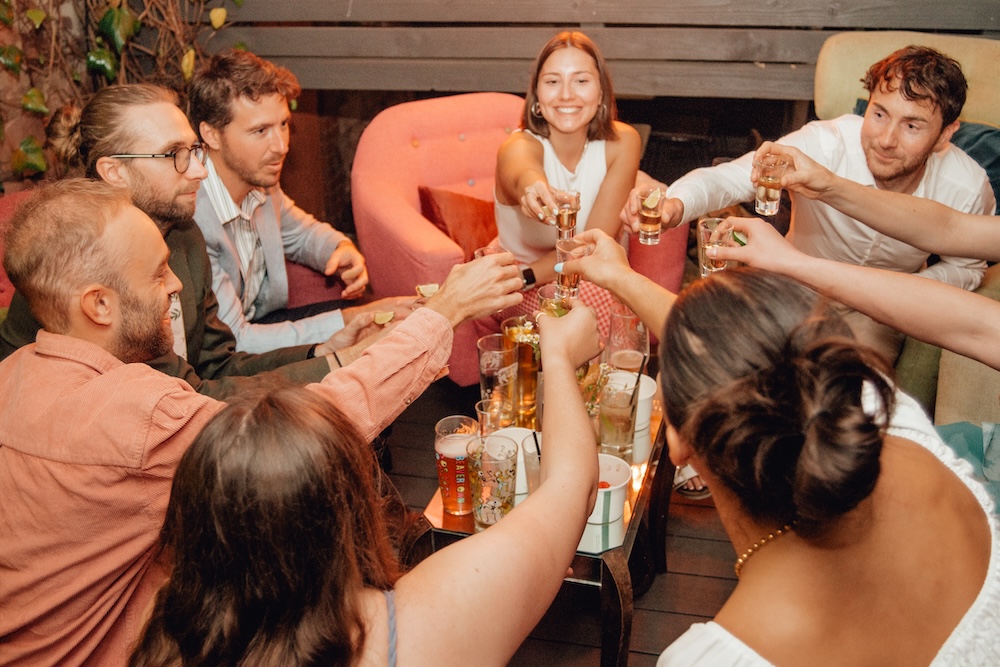 A group of friends raising glasses in a celebratory toast at The Square Club, one of the best party venues in Bristol. Seated in a cozy lounge area, the group of seven is smiling and engaging in the festive moment, holding up glasses of beer and shot glasses filled with clear liquid. The ambiance of the room reflects the social and energetic atmosphere perfect for any gathering at this Bristol-based venue.