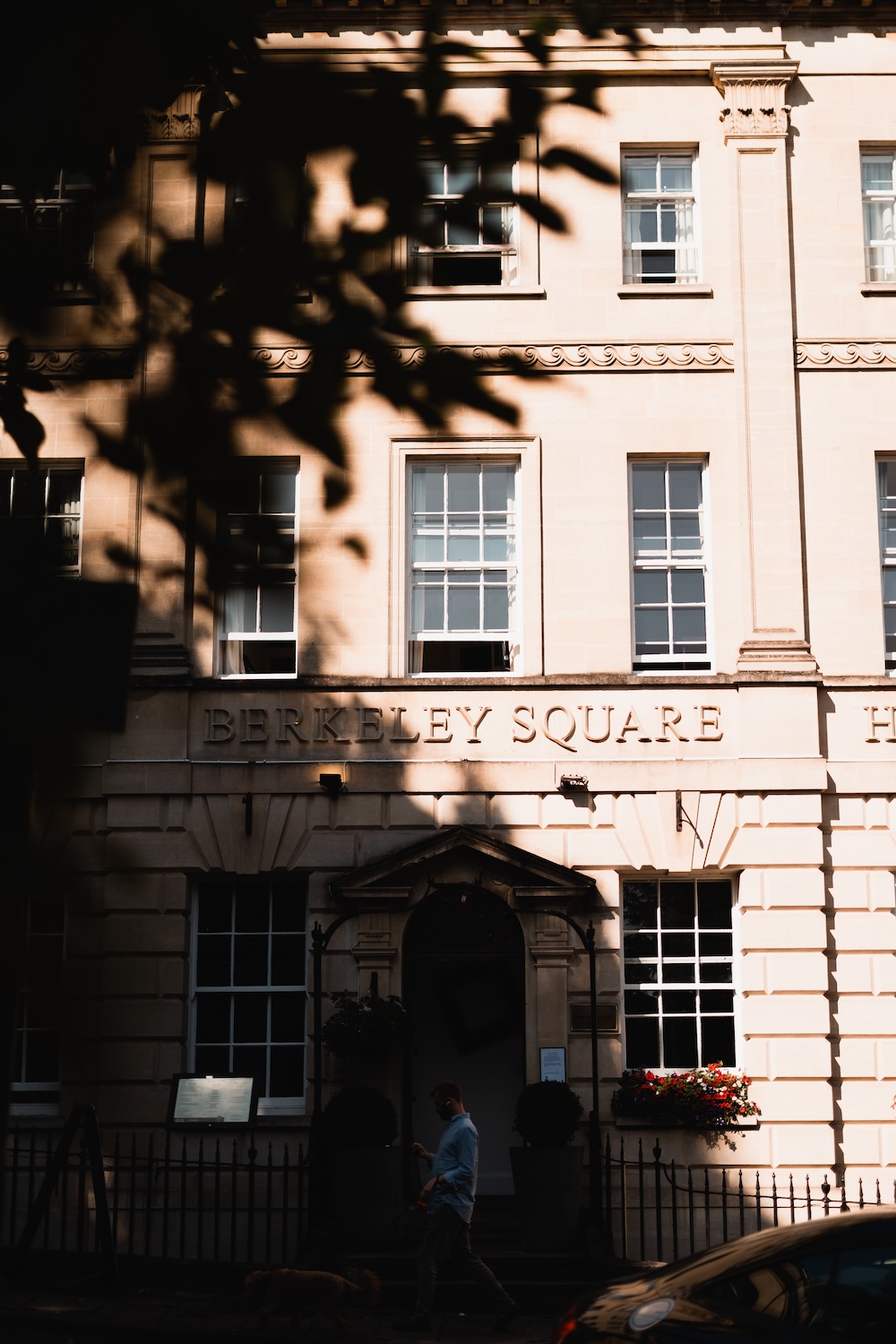 This image showcases the exterior of a historical building with "Berkeley Square" inscribed on its facade, a well-known wedding venue in Bristol. The building has a classic Georgian design, featuring tall windows and decorative moldings, bathed in soft sunlight. Shadows from nearby trees create an inviting, intimate atmosphere. The structure's grand entrance is visible, flanked by potted plants, and a person walks by with a dog, adding to the urban charm of this Bristol wedding venue. The setting exudes a timeless elegance, perfect for couples seeking an iconic location in the heart of the city.