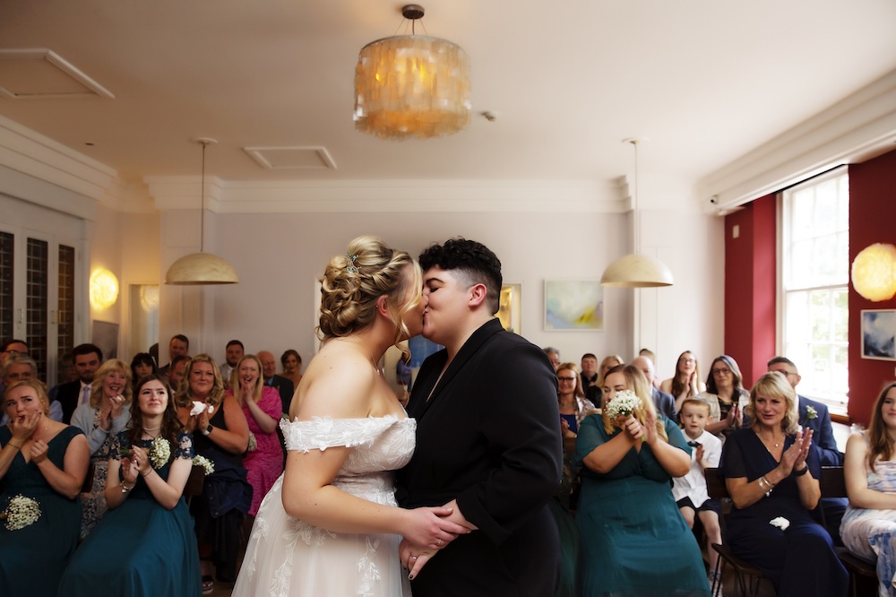 This photograph captures a heartwarming moment during a wedding ceremony inside a stylish room at a wedding venue in Bristol. A newlywed couple shares a kiss in front of their guests, creating a romantic and emotional atmosphere. The bride wears an off-shoulder wedding dress, while the other spouse is dressed in a chic black suit. Behind them, friends and family applaud and smile, seated in a light-filled space adorned with contemporary decor. This intimate wedding venue in Bristol provides an elegant setting for celebrating love, with its soft lighting, modern fixtures, and a layout perfect for close gatherings.