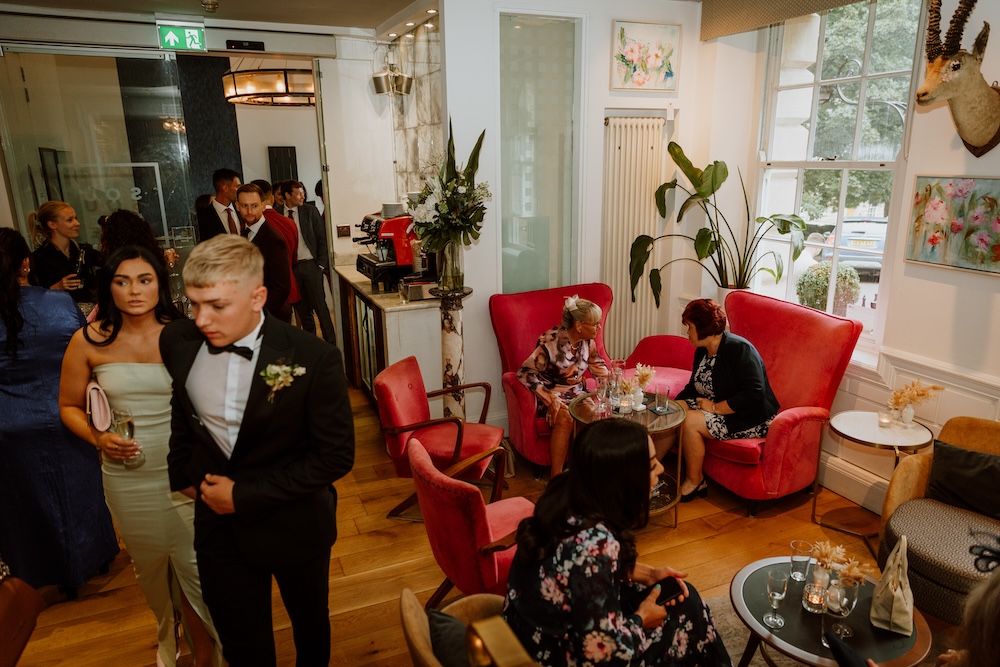 A lively cocktail hour at a wedding venue in Bristol is depicted in this image. Guests socialize in a cozy, chic lounge area with vibrant red armchairs and wooden floors, perfect for relaxing between ceremony and reception events. The venue's sophisticated decor, featuring floral artwork and stylish lighting, sets a modern yet intimate mood. A man in a black tuxedo and a woman in an elegant blue dress stand near the bar area, while others enjoy drinks and conversations. This versatile wedding venue in Bristol offers an ideal mix of comfort and class for couples looking for both style and substance.