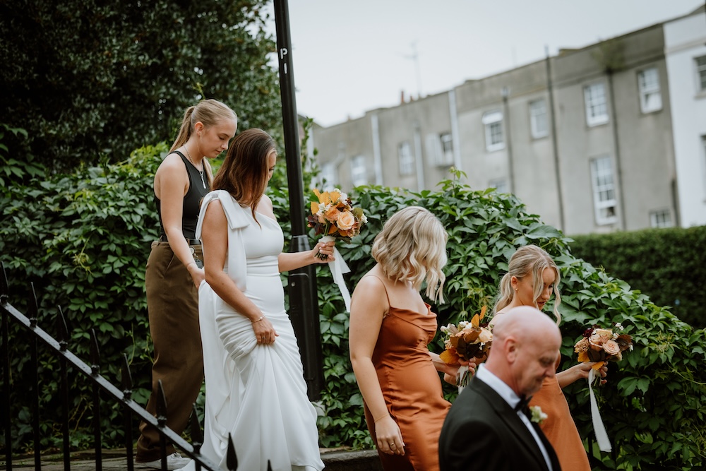 This image shows a bridal party walking down a staircase, with a backdrop of lush greenery outside a wedding venue in Bristol. The bride, wearing a sleek white gown, leads the group, followed by bridesmaids in stunning burnt orange dresses, each holding vibrant floral bouquets. The outdoor space, surrounded by ivy-covered walls, offers a serene, picturesque setting for wedding photography. This Bristol wedding venue provides a blend of natural beauty and urban charm, creating an ideal spot for capturing unforgettable moments.