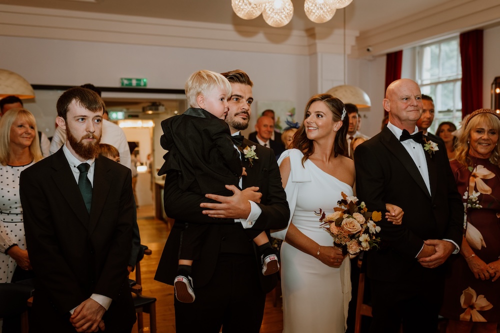 A touching scene during a wedding ceremony at a stylish indoor venue in Bristol is displayed in this image. The bride, wearing an elegant one-shoulder gown, stands beside her groom, who holds their young child in his arms. They are surrounded by family and friends, with smiles and tender expressions on their faces. The intimate wedding venue in Bristol is designed with modern lighting, soft tones, and minimalist decor, providing a serene and comfortable environment for the couple and their guests.