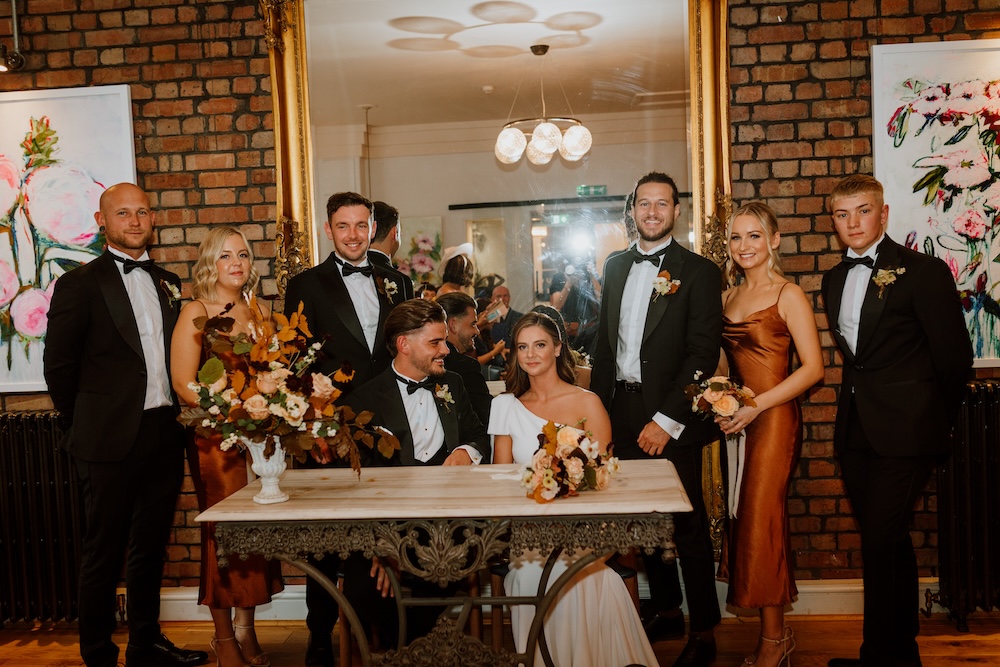 This formal wedding portrait features the bride and groom seated at a marble table, flanked by their bridal party, at a sophisticated wedding venue in Bristol. The wedding party, dressed in sharp black tuxedos and stylish orange bridesmaid dresses, stands in front of a brick wall adorned with contemporary floral artwork. The venue's combination of exposed brick and modern decor creates a trendy, fashionable setting for wedding celebrations, appealing to couples seeking a chic yet cozy Bristol wedding venue.