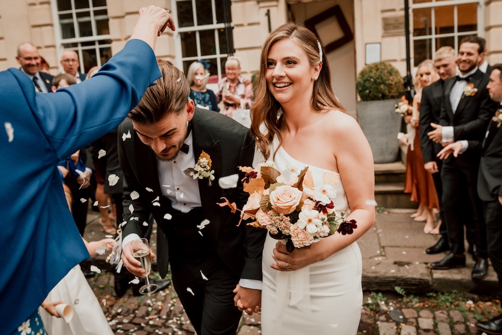 Guests gather outside a wedding venue in Bristol as the newlyweds walk through a shower of confetti. The bride, holding a bouquet of autumn-inspired flowers, smiles radiantly, while the groom ducks playfully under the falling confetti. The venue’s classical architecture, with large windows and well-maintained stonework, provides a stunning backdrop for this joyful moment. The Bristol wedding venue’s elegant exterior adds a sense of grandeur to the couple’s exit, making it a memorable part of the day.