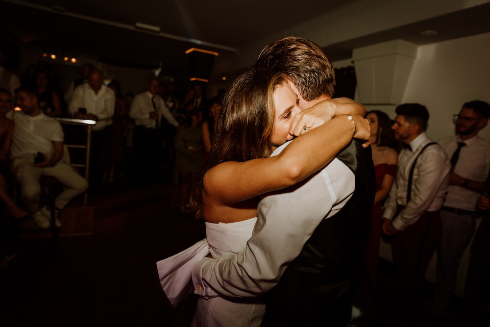 This image captures the emotional embrace of a newlywed couple during their first dance at a wedding venue in Bristol. The bride wraps her arms tightly around the groom, expressing pure joy and love in this candid moment. The venue’s softly lit dance floor, surrounded by friends and family, provides an intimate space for this heartfelt moment. The wedding venue in Bristol features modern lighting, a spacious dance area, and a warm ambiance, making it an ideal choice for couples seeking a venue that offers both elegance and intimacy.