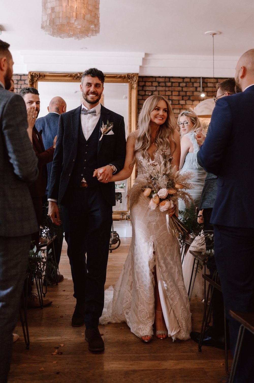 In this image, the bride and groom are walking down the aisle of an indoor wedding venue in Bristol. The groom is dressed in a sharp navy blue suit paired with a bowtie, while the bride stuns in a form-fitting lace gown, holding a rustic bouquet of dried flowers and peach-colored roses. The venue features warm, wooden floors and modern hanging lights, giving it an elegant yet intimate atmosphere. Guests line the aisle, smiling and congratulating the couple. This Bristol wedding venue blends a touch of tradition with contemporary design, offering a perfect backdrop for a chic and romantic ceremony.