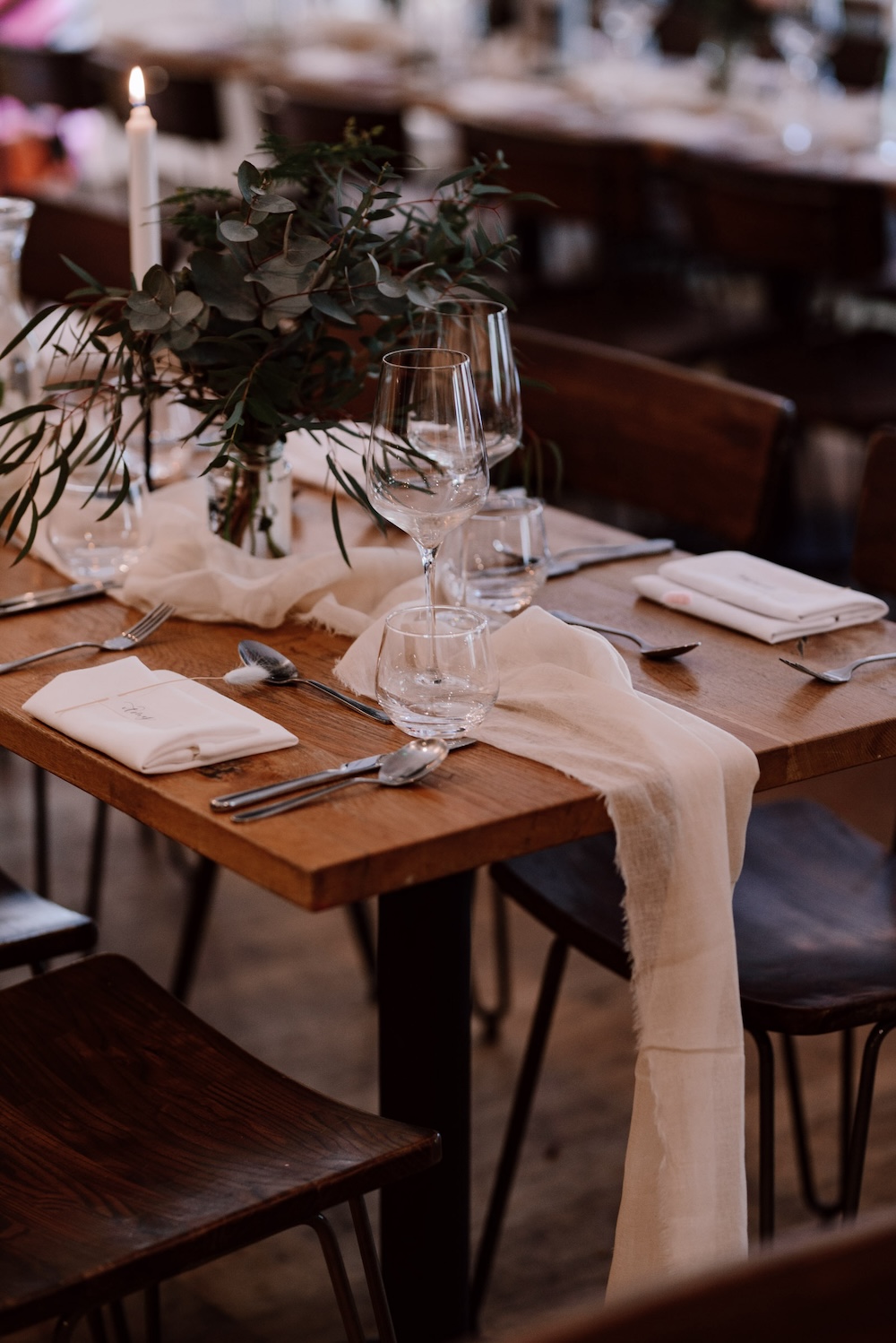 This close-up image highlights an elegantly set table at a wedding reception, located at a wedding venue in Bristol. The wooden table features delicate glassware, silver utensils, and minimalist floral arrangements with greenery. A single lit candle adds a warm glow, while soft white fabric drapes over the table, adding a touch of elegance. The rustic yet refined aesthetic of this Bristol wedding venue makes it ideal for couples seeking a cozy yet sophisticated setting for their reception dinner.