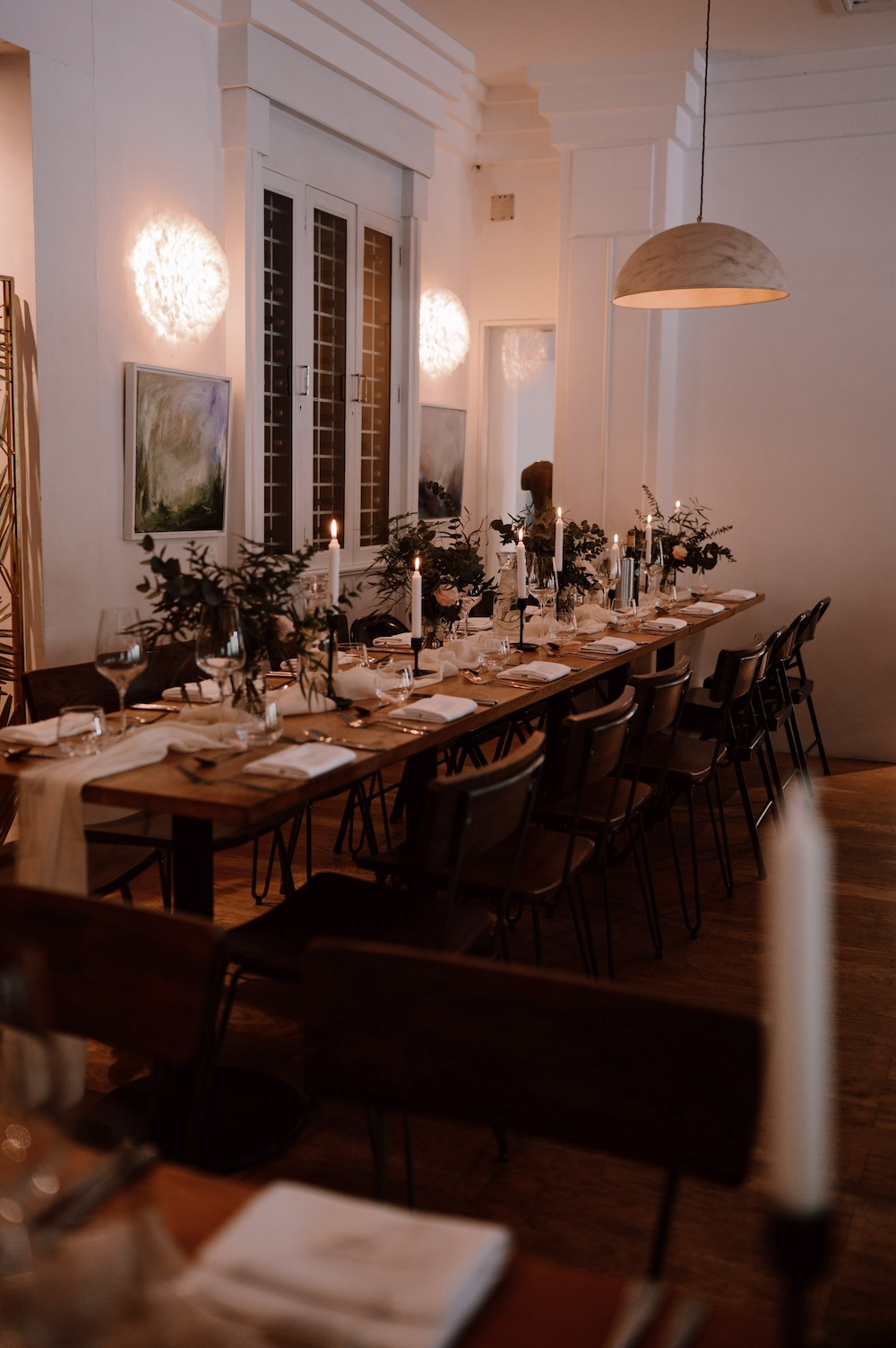 The dining area at this wedding venue in Bristol is styled with long wooden tables adorned with simple yet elegant greenery, candles, and glassware. The space is softly lit by modern hanging lamps, creating an intimate and welcoming environment for the guests. The neutral color palette and minimalist decor add to the serene ambiance of the venue, which is perfect for a relaxed yet refined wedding reception. This wedding venue in Bristol offers a contemporary design that complements the couple's personalized touches.