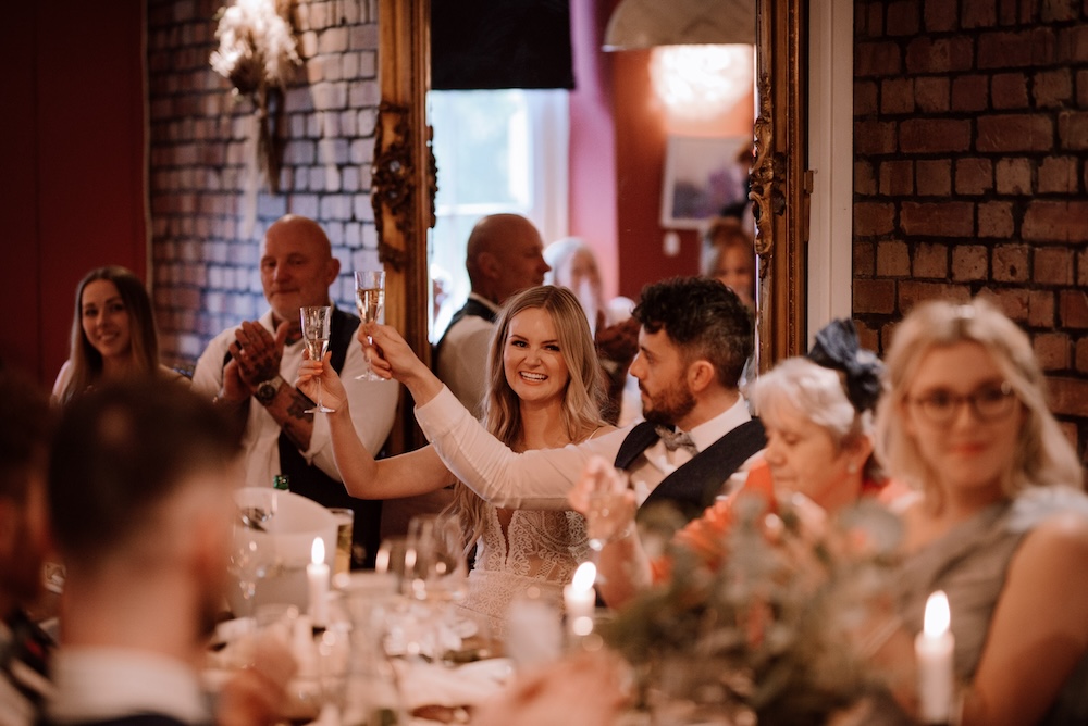 The bride and groom are seated at the head table during their wedding reception at a Bristol wedding venue, raising their glasses in a celebratory toast. The bride smiles brightly, wearing a stunning lace dress, while the groom looks on proudly. Guests surrounding them also join in the toast, adding to the festive and warm atmosphere. The venue, with its exposed brick walls and soft lighting, creates a cozy yet stylish setting for the couple's special day. This wedding venue in Bristol provides the perfect combination of intimate charm and modern décor for an unforgettable celebration.