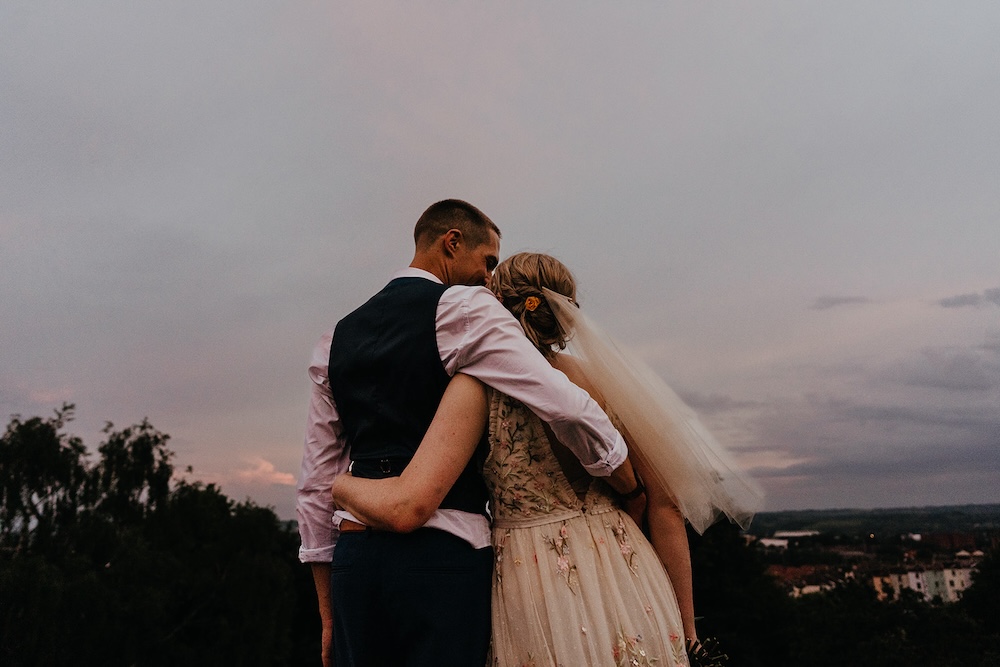 This beautifully candid image shows a couple embracing outdoors at sunset, with a dramatic sky in the background. The bride, wearing a gown with floral details, leans into the groom as they share a quiet, intimate moment. The soft evening light casts a romantic glow over them, and the view of the city beyond adds a unique touch to the image. This Bristol wedding venue is ideal for couples seeking both picturesque outdoor settings and proximity to the city, allowing for a blend of natural beauty and urban elegance.