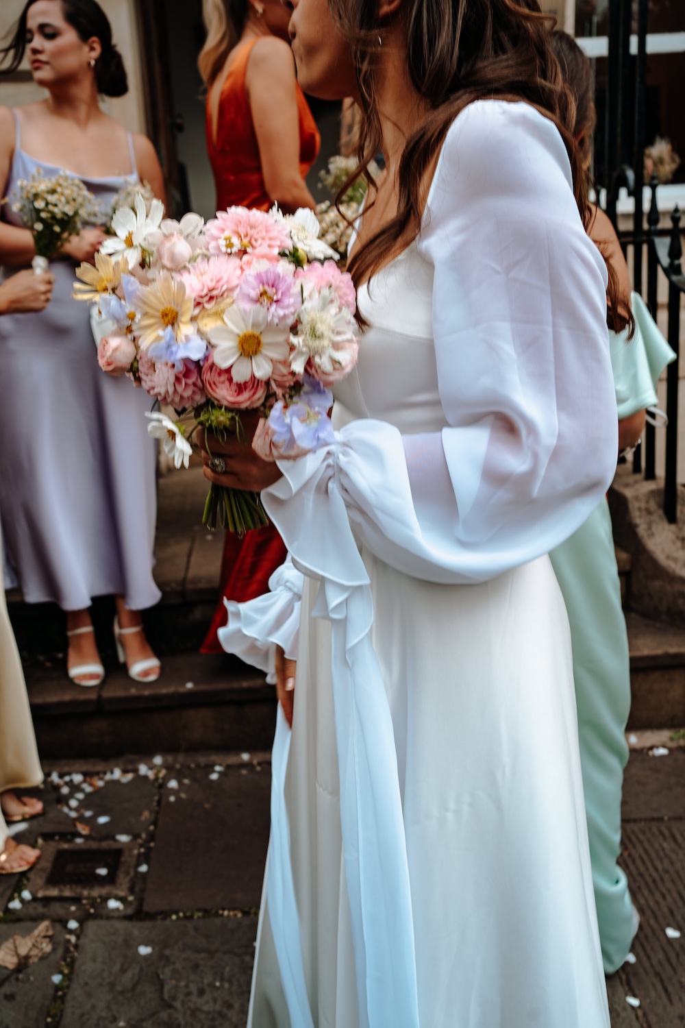 This image focuses on the bride’s bouquet at a wedding venue in Bristol, filled with soft pink and white flowers, including delicate daisies and peonies. The bride, dressed in a white gown with billowing sleeves, stands alongside her bridesmaids, whose dresses add pops of color to the scene. The close-up of the bouquet, combined with the elegant simplicity of the wedding attire, emphasizes the beauty and attention to detail at The Square Club Bristol wedding venue, where every element contributes to a romantic, floral-themed celebration.