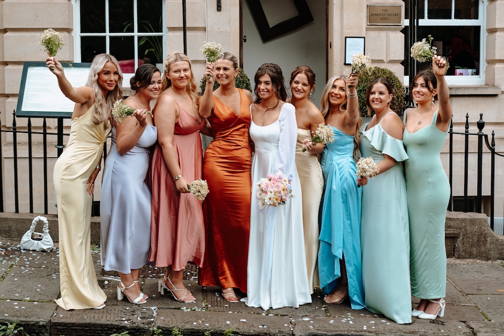 This image showcases a joyful bridal party standing in front of The Square Club, a wedding venue in Bristol. The bride is surrounded by her bridesmaids, each wearing colorful dresses in shades of yellow, pink, green, and blue. The women are all smiling and holding small bouquets of flowers, celebrating outside the historic facade of the venue. The Square Club's elegant stonework and large windows provide a timeless backdrop, making it an ideal setting for wedding photography that captures both the vibrancy of the bridal party and the classic architecture of this Bristol wedding venue.