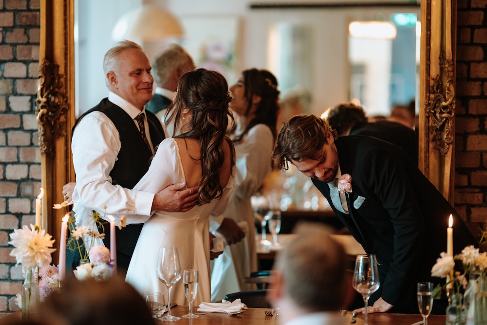 A heartwarming moment unfolds as the bride shares a dance with an older guest, possibly her father, while the groom stands beside them in admiration. This indoor scene takes place at The Square Club, a well-known wedding venue in Bristol. The reflection in the large ornate mirror, the elegant brick walls, and the romantic lighting all contribute to the venue’s warm and classic ambiance. The Square Club offers a refined and intimate setting for weddings, where family moments like these are beautifully framed by the venue’s stylish interior.