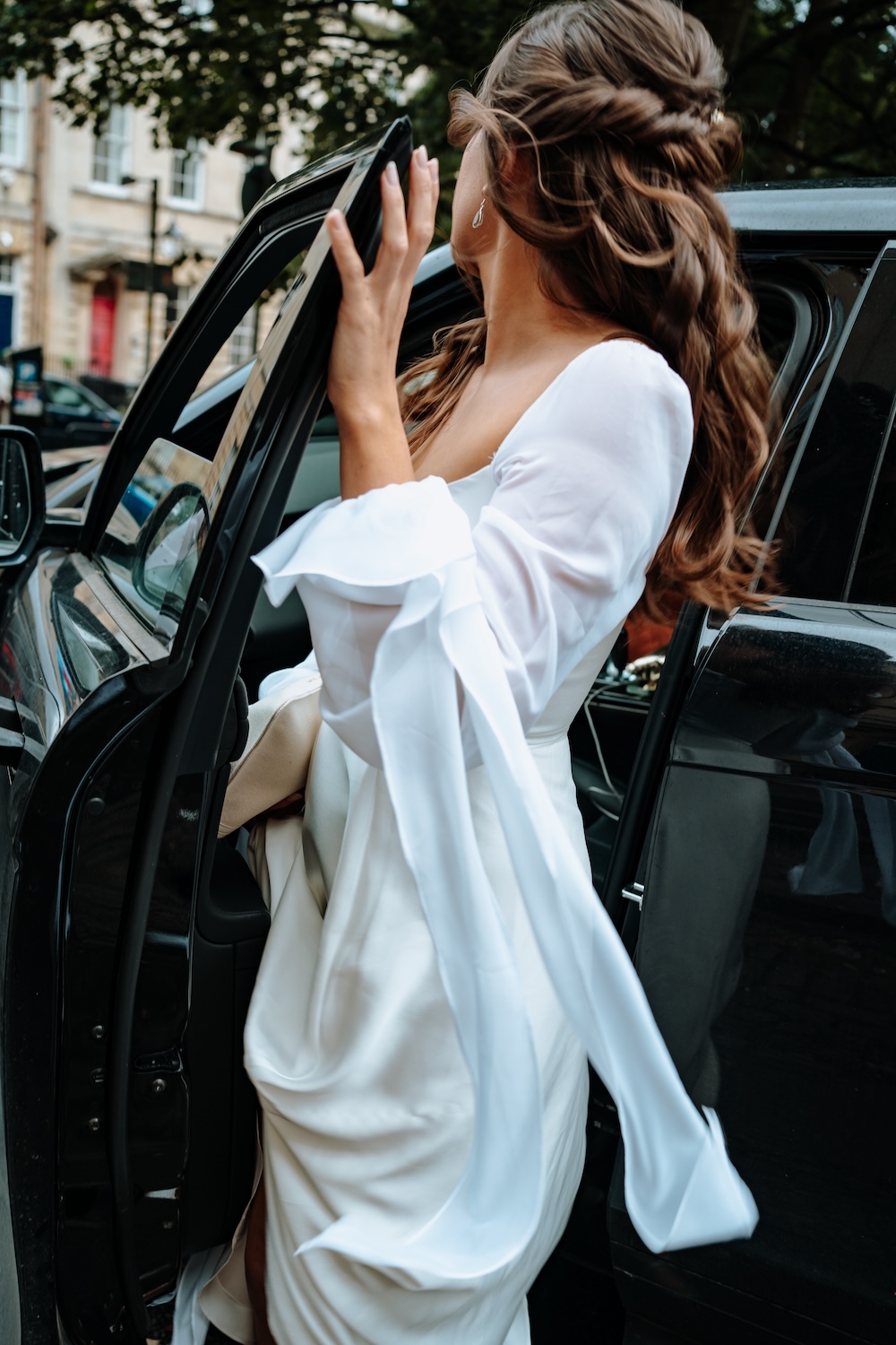 The bride, wearing an ethereal long-sleeved gown, is captured entering a car parked outside The Square Club, a beautiful wedding venue in Bristol. Her intricate hairstyle and flowing gown complement the classic architecture of the surrounding streets. This shot, taken during a moment of transition, reflects the sophisticated yet personal experience that The Square Club offers to couples on their wedding day. This Bristol wedding venue blends modern city life with timeless romance, perfect for an unforgettable event.