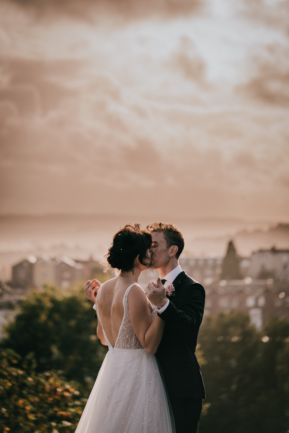 The bride and groom share a romantic kiss atop a hill with the city of Bristol in the background. This outdoor shot, taken at The Square Club, showcases the venue’s stunning location, where couples can enjoy picturesque views of the surrounding cityscape. The soft evening light enhances the romantic mood of this wedding venue in Bristol, making it an ideal spot for unforgettable moments like this one. The bride’s backless gown and the groom’s formal attire are perfectly complemented by the natural beauty and elegance of the setting.