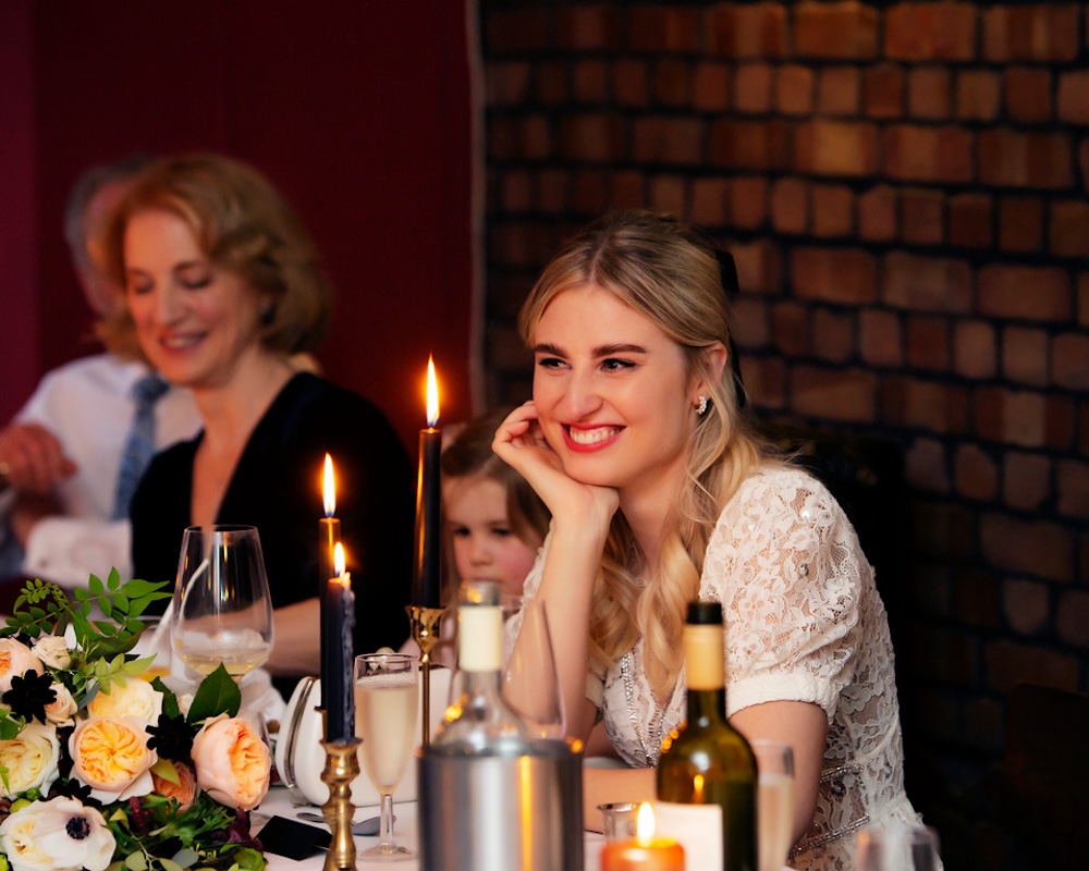 This intimate reception scene captures the bride smiling warmly as she listens to wedding speeches during her reception at The Square Club, a stylish wedding venue in Bristol. The bride is seated at a beautifully decorated table, surrounded by soft candlelight and fresh floral arrangements. The venue’s exposed brick walls and ambient lighting create a cozy, intimate atmosphere. The Square Club offers a perfect blend of modern elegance and classic charm, making it an excellent choice for couples seeking a romantic and inviting space for their wedding in Bristol.