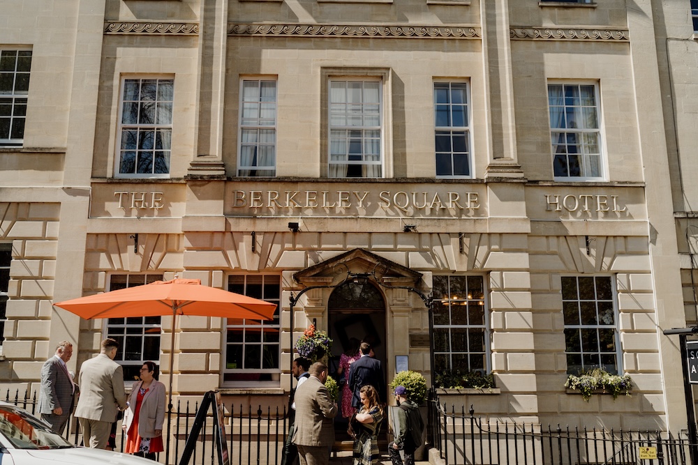 A detailed exterior shot of The Square Club, a popular wedding venue in Bristol, showcases its historic and elegant architecture. The sunlight highlights the grand entrance of The Berkeley Square Hotel, where wedding guests are gathered beneath a bright orange umbrella. The iconic stone facade and large windows of this Bristol wedding venue create an inviting yet sophisticated ambiance, making it a sought-after location for stylish city weddings.