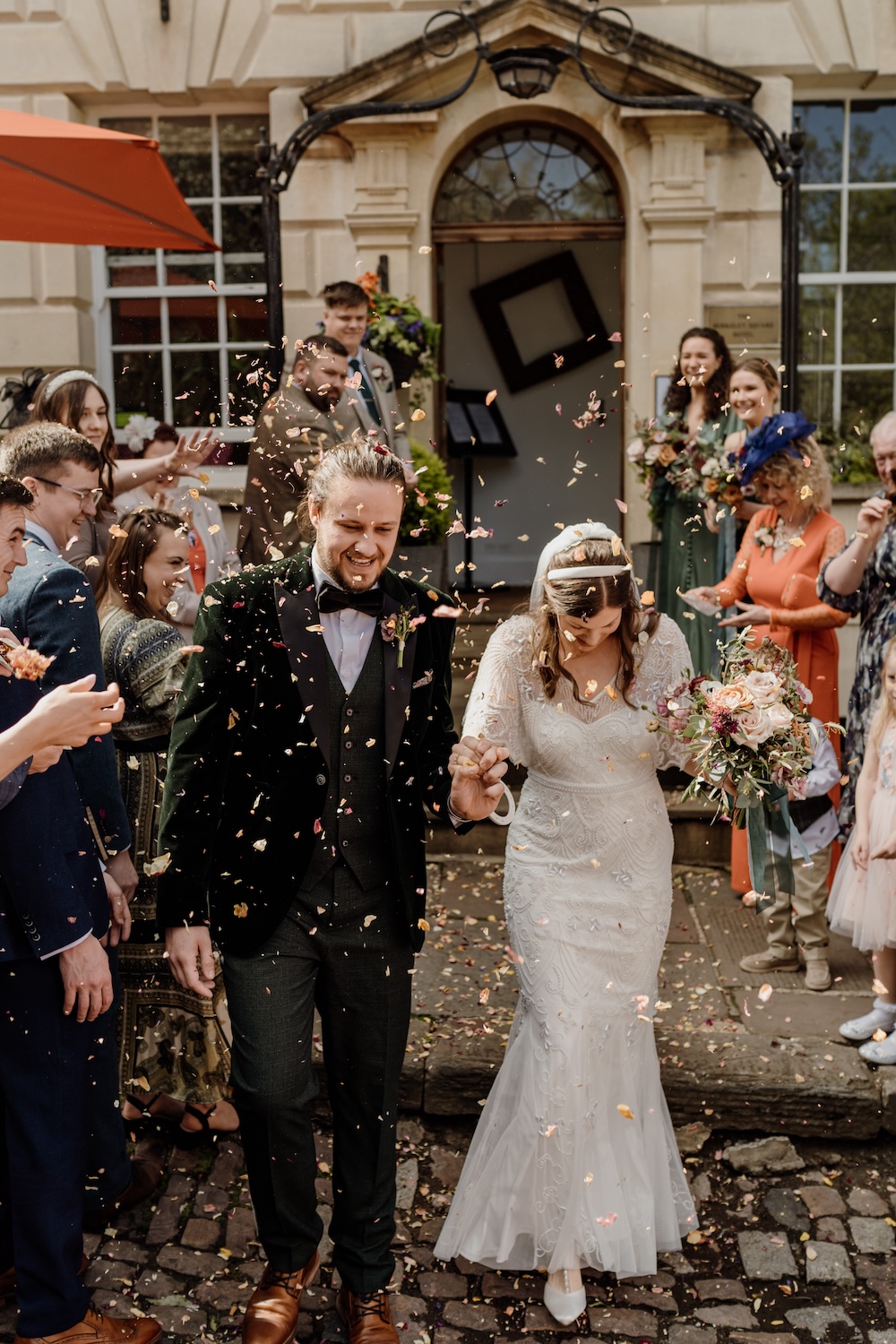 A bride and groom walk hand-in-hand under a cascade of confetti outside The Square Club, a stylish wedding venue in Bristol. Surrounded by smiling guests, the newlyweds beam with happiness as the celebratory moment unfolds. The venue’s elegant exterior, featuring classic stone architecture, provides the perfect backdrop for such moments of joy and connection, making The Square Club a standout choice for a romantic city wedding in Bristol.